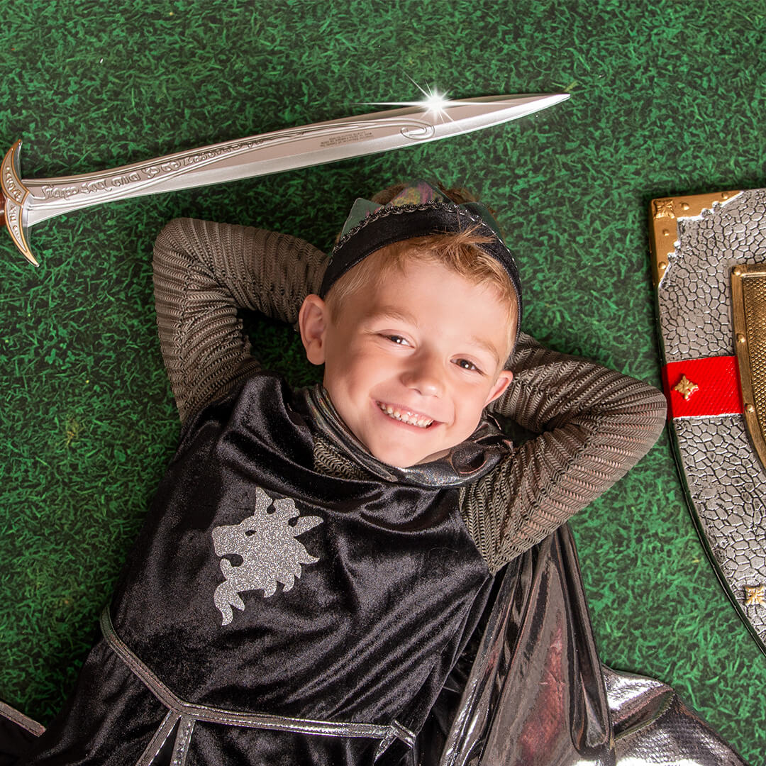 Boy dressed in a knights costume posing on the floor with a sword and shield prop.