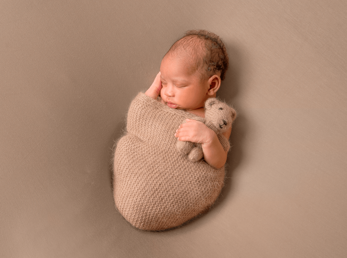 Newborn photographed on a brown blanket, holding a teddy bear.