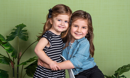 Sisters smiling for professional children pictures on Willow background at JCPenney Portraits.